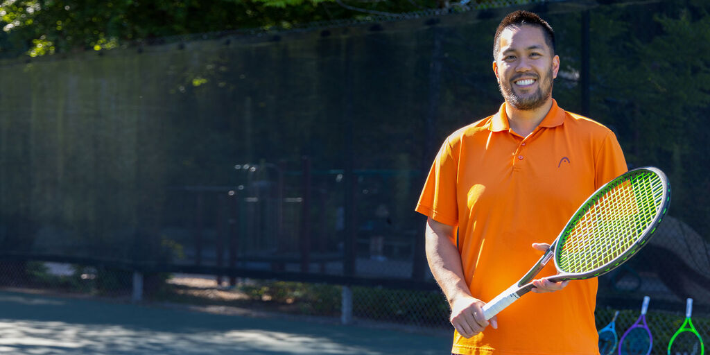 man outside in an orange Head t-shirt holding a tennis racquet and smiling